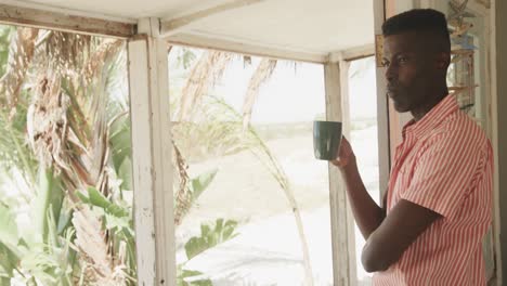 Nachdenklicher-Afroamerikanischer-Mann,-Der-Kaffee-Trinkt-Und-Sich-In-Der-Tür-Eines-Strandhauses-Lehnt,-Zeitlupe