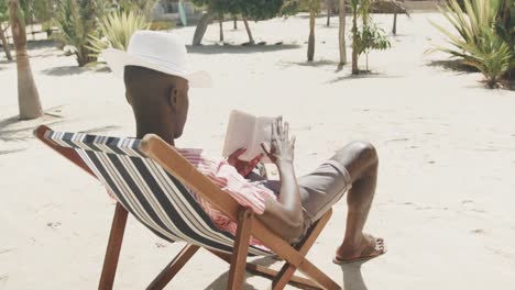 Hombre-Afroamericano-Relajado-Sentado-En-Una-Tumbona-Leyendo-Un-Libro-En-Una-Playa-Soleada,-Cámara-Lenta