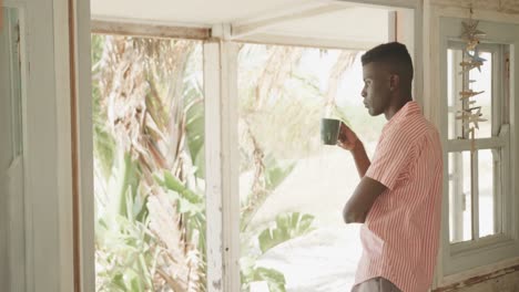 Thoughtful-african-american-man-drinking-coffee-leaning-in-doorway-of-beach-house,-slow-motion