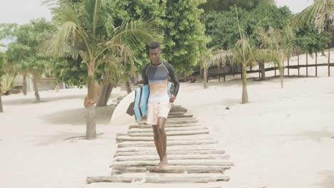 Happy-african-american-man-carrying-surfboard-running-on-boardwalk-on-sunny-beach,-slow-motion