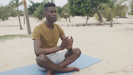 Hombre-Afroamericano-Relajado-Haciendo-Yoga,-Sentado-En-Una-Alfombra-Meditando-En-La-Playa,-Cámara-Lenta