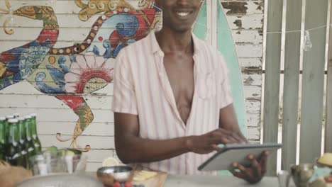 Retrato-De-Un-Feliz-Barman-Afroamericano-Usando-Una-Tableta-Detrás-Del-Mostrador-En-El-Bar-De-La-Playa,-Cámara-Lenta