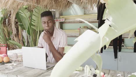 Happy-african-american-man-using-laptop-behind-the-counter-of-surf-rental-beach-shack,-slow-motion