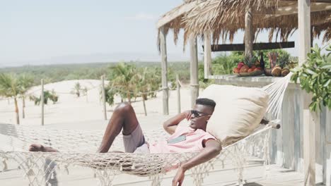 Happy-african-american-man-lying-in-hammock-wearing-sunglasses-on-sunny-beach,-slow-motion