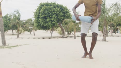 Happy-african-american-man-rolling-up-mat-after-doing-yoga-at-the-beach,-slow-motion