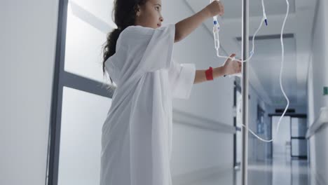 Happy-biracial-sick-girl-patient-holding-drip-in-hospital-corridor-in-slow-motion