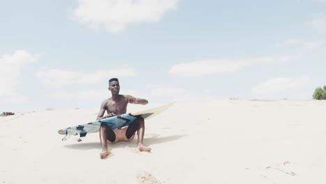Happy-african-american-man-holding-surfboard,-getting-up-from-sitting-on-sunny-beach,-slow-motion