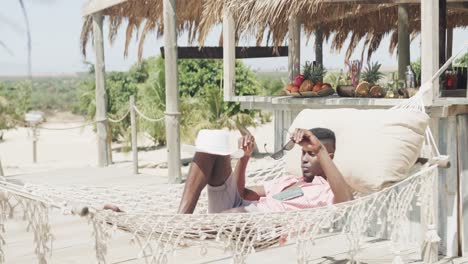 Happy-african-american-man-lying-in-hammock-putting-hat-over-face-on-sunny-beach,-slow-motion