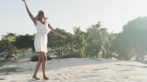 Happy-biracial-woman-in-sundress-and-sunhat-dancing-on-sunny-beach,-slow-motion