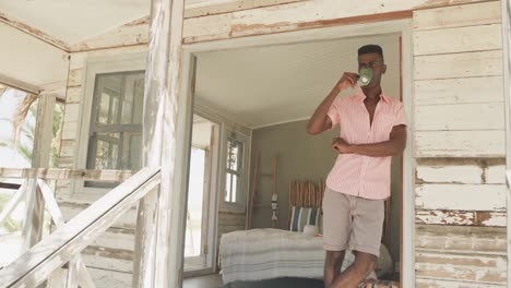 African-american-man-drinking-coffee-on-the-porch-of-wooden-beach-house-in-the-sun,-slow-motion