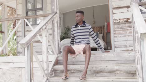 African-american-man-sitting-on-front-steps-of-wooden-beach-house-relaxing-in-the-sun,-slow-motion