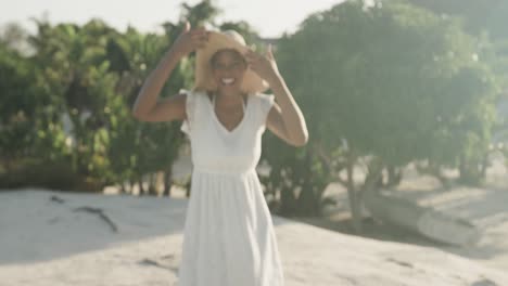 Portrait-of-happy-biracial-woman-in-sundress-and-sunhat-dancing-on-sunny-beach,-slow-motion