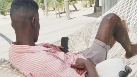 Afroamerikanischer-Mann-Liegt-In-Der-Hängematte-Und-Benutzt-Smartphone-Am-Sonnigen-Strand,-Zeitlupe
