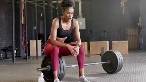Müde-Afroamerikanische-Frau-Sitzt-Auf-Der-Langhantel-Und-Ruht-Sich-Nach-Dem-Gewichtheben-Im-Fitnessstudio-Aus,-Zeitlupe