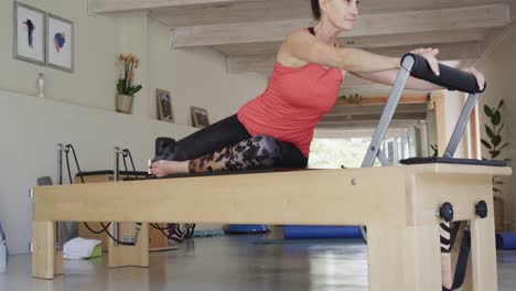 Fit-senior-caucasian-woman-stretching-on-reformer-in-pilates-studio,-unaltered,-in-slow-motion