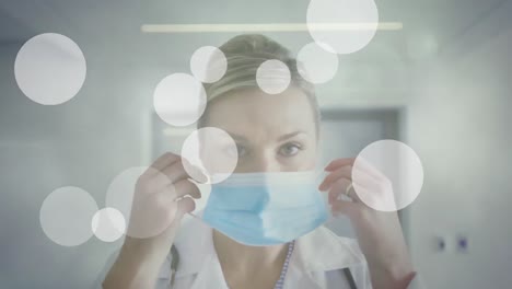 Animation-of-white-spots-over-caucasian-female-doctor-wearing-a-face-mask-at-hospital