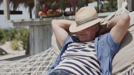 Happy-senior-caucasian-man-in-hat-relaxing-in-hammock-in-the-sun-by-beach-bar,-in-slow-motion