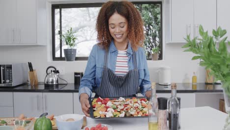 Portrait-of-happy-biracial-woman-in-apron-holding-seasoned-vegetables-on-baking-tray,-slow-motion