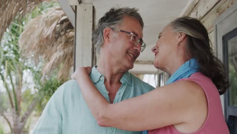 Happy-senior-caucasian-couple-talking-and-embracing-on-porch-of-beach-house,-in-slow-motion