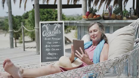 Happy-senior-caucasian-woman-lying-in-hammock-using-tablet-by-beach-bar,-in-slow-motion