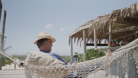 Relaxed-senior-caucasian-man-sitting-in-hammock-using-tablet-by-beach-bar,-in-slow-motion