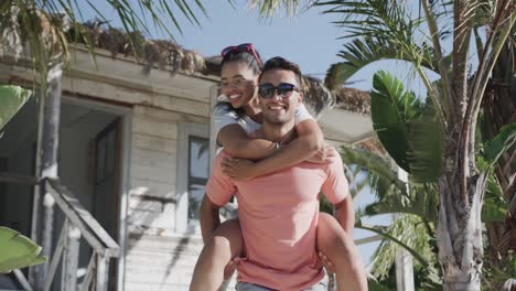 Happy-diverse-couple-having-fun-piggybacking-outside-wooden-beach-house-in-the-sun,-in-slow-motion