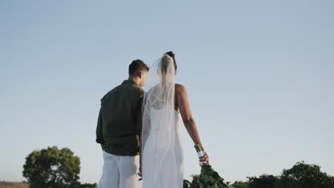 Rear-view-of-diverse-bride-and-groom-walking-away-holding-hands-at-beach-wedding,-in-slow-motion