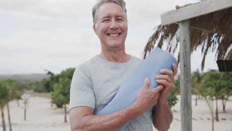 Portrait-of-happy-senior-caucasian-man-holding-yoga-mat-and-smiling-on-beach,-in-slow-motion