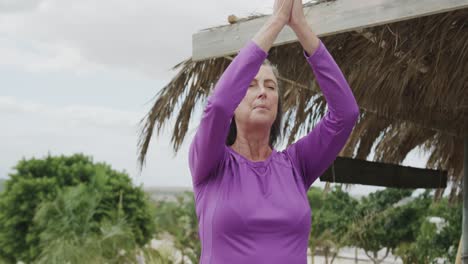 Happy-senior-caucasian-woman-practicing-yoga-meditation-on-beach,-in-slow-motion
