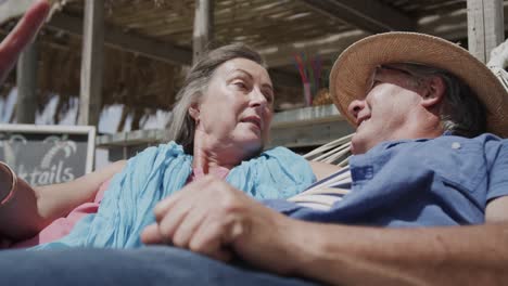 Happy-senior-caucasian-couple-lying-on-hammock-talking-outside-beach-bar,-in-slow-motion