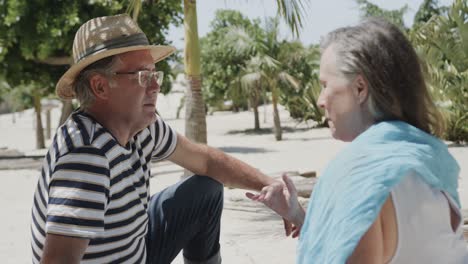 Happy-senior-caucasian-couple-sitting-on-beach-talking-in-the-sun,-in-slow-motion