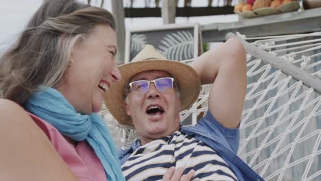 Laughing-senior-caucasian-couple-relaxing-on-hammock-outside-beach-house,-in-slow-motion