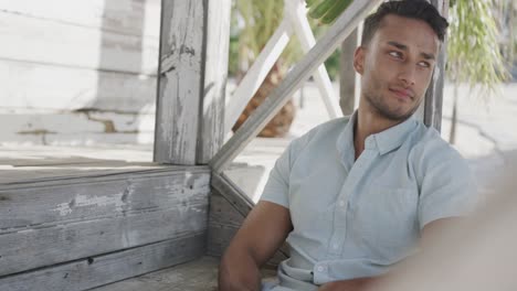 Thoughtful-biracial-man-using-smartphone-sitting-on-porch-of-wooden-beach-house,-in-slow-motion