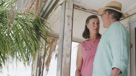 Happy-senior-caucasian-couple-talking-on-porch-of-wooden-beach-house-enjoying-view,-in-slow-motion