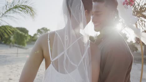 Happy-diverse-bride-and-groom-touching-heads-and-smiling-at-beach-wedding,-in-slow-motion