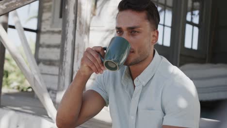 Thoughtful-biracial-man-drinking-coffee-sitting-on-porch-of-beach-house,-in-slow-motion