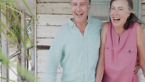 Portrait-of-happy-senior-caucasian-couple-laughing-standing-on-porch-of-beach-house,-in-slow-motion