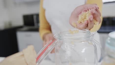 Midsection-of-biracial-woman-pouring-pasta-from-hand-into-storage-jar-in-kitchen,-in-slow-motion