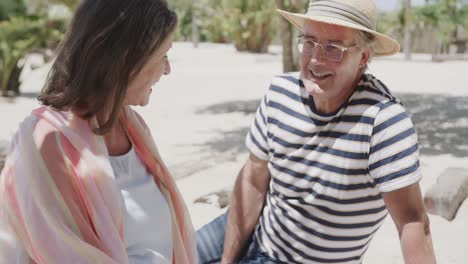 Happy-senior-caucasian-couple-sitting-on-beach-talking-in-the-sun,-in-slow-motion