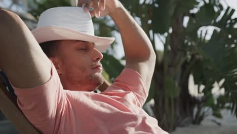 Relaxed-biracial-man-sitting-in-sun-on-beach-putting-hat-over-face,-in-slow-motion