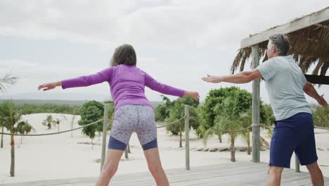 Happy-senior-caucasian-couple-practicing-yoga-standing-on-beach-sun-deck,-in-slow-motion