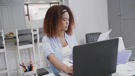 Busy-biracial-woman-working-on-laptop-and-inspecting-documents-at-home,-in-slow-motion