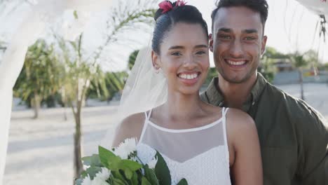 Portrait-of-happy-diverse-bride-and-groom-smiling-at-sunny-beach-wedding,-in-slow-motion