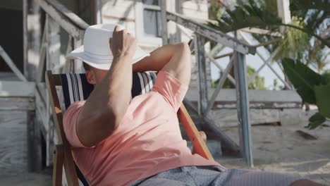 Relaxed-biracial-man-sitting-in-sun-with-hat-over-face-outside-beach-house,-in-slow-motion