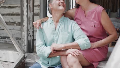 Portrait-of-happy-senior-caucasian-couple-embracing-sitting-on-porch-of-beach-house,-in-slow-motion