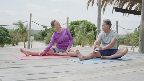 Happy-senior-caucasian-couple-practicing-yoga-sitting-on-beach-sun-deck,-in-slow-motion