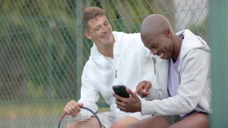 Fröhliche,-Vielfältige-Männliche-Freunde,-Die-Auf-Ihr-Smartphone-Schauen-Und-Auf-Dem-Tennisplatz-Lachen,-Zeitlupe