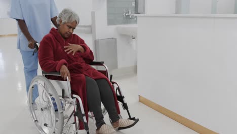 Diverse-female-doctor-pushing-senior-female-patient-in-wheelchair-in-hospital-corridor,-slow-motion