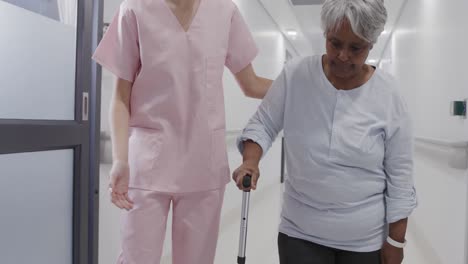 Diverse-female-doctor-talking-with-senior-female-patient-walking-with-stick-in-corridor,-slow-motion
