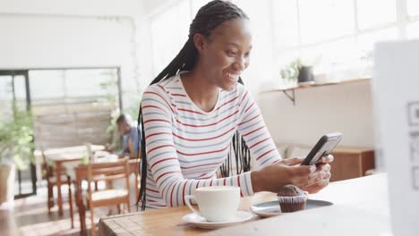 Fröhliche-Afroamerikanische-Frau-Mit-Smartphone-Am-Tisch-Im-Sonnigen-Café-Sitzend,-Zeitlupe